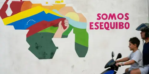 Mural przedstawiający mapę Wenezueli z flagą i hasłem "Somos Esequibo". Dwóch chłopców na skuterze przejeżdża obok.