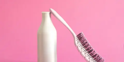 Eine weiße Flasche, die wie ein **collagen shampoo** aussieht, lehnt an einer Haarbürste auf rosa Hintergrund.