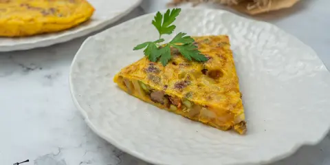 Un trozo de tortilla paisana, con patatas, verduras y chorizo, servida en un plato blanco texturizado.
