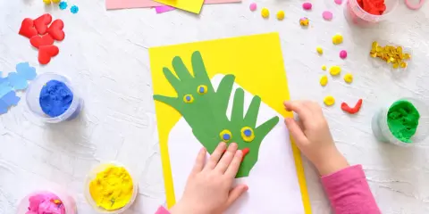 Kreatives Basteln mit Kleinkindern: Hände werden zu grünen Drachen auf gelbem Papier, umgeben von bunter Knete und Bastelmaterialien.