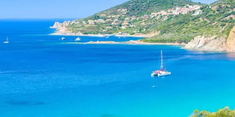 Un catamaran blanc avec des accents rouges navigue sur des eaux bleues cristallines, près d'une côte verdoyante et d'un village pittoresque.