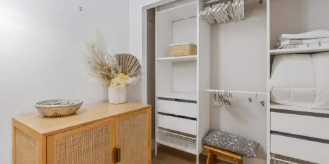 Un armario blanco organizado, listo para limpiar armarios por dentro. Cestas, ropa de cama y un jarrón con flores secas decoran el espacio.