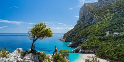 Escursionista ammira una baia cristallina con una spiaggia bianca, incastonata tra imponenti scogliere verdi. Un paesaggio mozzafiato che ricorda la bellezza della montagna centro Italia.