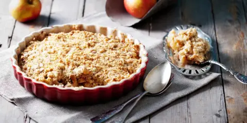 Un délicieux crumble pomme individuel, doré et croustillant, prêt à être dégusté. Des pommes fraîches sont visibles en arrière-plan.