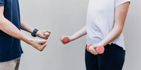 Dwie osoby ćwiczą z hantlami. Jedna osoba pokazuje zaciśniętą pięść, druga trzyma różowe hantle. Z czym do fizjoterapeuty?