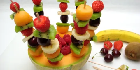 Une **brochette de fruits** colorée avec fraises, kiwis, bananes, cerises et melon, présentée dans une moitié de melon.