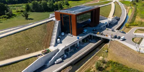 Nowoczesny budynek muzeum bitwy warszawskiej z cortenowską fasadą, otoczony zielenią i betonowymi konstrukcjami.