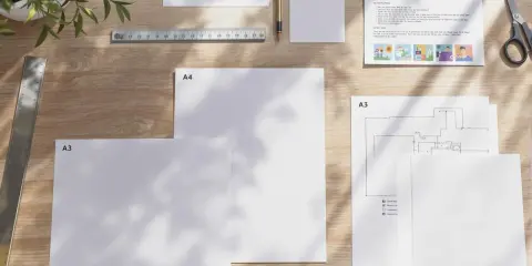 Na drewnianym biurku leżą kartki papieru w formatach A4 i A3, linijka, ołówek i nożyczki. Jedna z kartek ma nadrukowany tekst i ilustracje, przypominając materiały do druku w rozmiarze papieru letter.