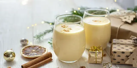 Zwei Gläser cremiger Cocktail mit Eigelb, garniert mit Zimt, umgeben von Weihnachtsdekorationen und Geschenken.