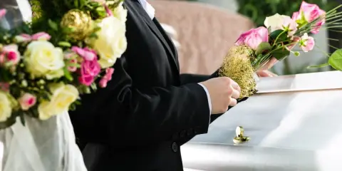 Osoba w czarnym garniturze kładzie bukiet róż na białej trumnie.