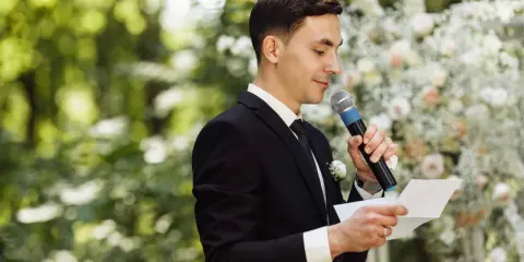 El maestro de ceremonias, con traje negro y micrófono, lee un discurso frente a un arco de flores.
