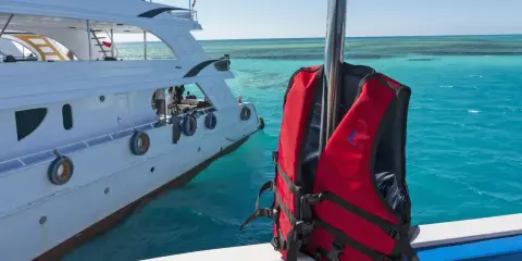 Gilet de sauvetage rouge prêt pour l'aventure en mer, à côté d'un yacht blanc. Les **œuvres mortes du bateau** se reflètent dans l'eau turquoise.