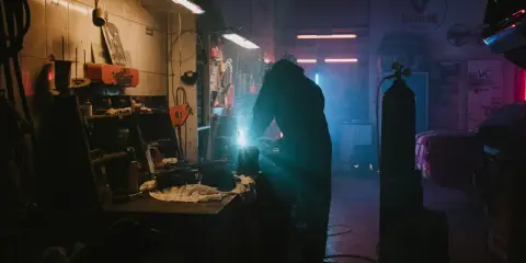 Un soudeur travaille dans un garage sombre, éclairé par des néons. La question "dans quel sens installer un néon dans un garage" plane dans l'air.