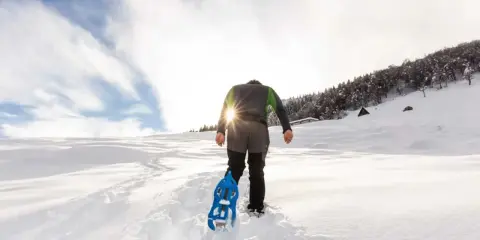 Uomo con ciaspole cammina su sentiero innevato, ideale per chi cerca dove ciaspolare in Piemonte. Sole splendente tra le nuvole.
