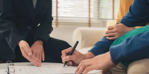 Podpisanie umowy najmu. Czy mogę wynająć mieszkanie rodziców? Dwie osoby siedzą przy stole, jedna podpisuje dokument, druga wskazuje na niego.