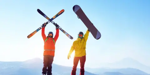 Dwóch narciarzy na szczycie góry, jeden z nartami, drugi z deską snowboardową, cieszy się słońcem i widokiem.