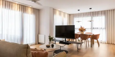 Acogedor salón de 20 metros cuadrados con sofá, mesa de centro, TV, y comedor. Cortinas blancas y suelo de madera crean un ambiente luminoso.