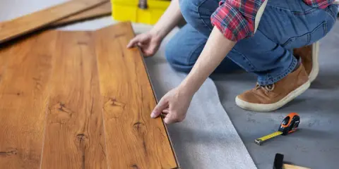 Persona instala tarima flotante, uniendo tablas de madera con cuidado. Herramientas como martillo y cinta métrica a mano.