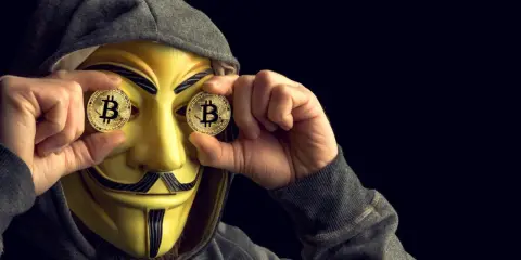 Osoba w masce Guya Fawkesa trzyma monety z Bitcoinem przed oczami. Czy MoonPay jest bezpieczny?