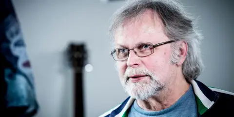 Clark Olofsson idag, med grått hår, skägg och glasögon, blickar mot betraktaren.