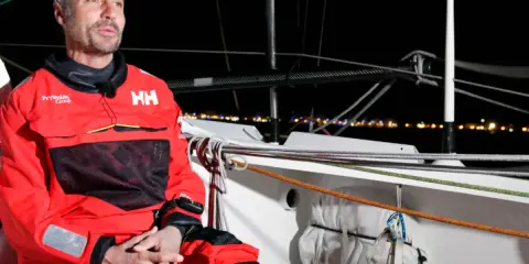 Giancarlo Pedote, skipper en tenue de voile rouge, sur un bateau la nuit, avec des lumières de ville en arrière-plan.