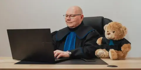 Prawnik w todze z pluszowym misiem obok, pracujący przy laptopie. Zastanawiasz się, ile kosztuje porada prawna?