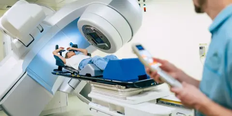 Ragazza sdraiata su lettino per radioterapia, tecnico in primo piano controlla il macchinario. Speranza di guarigione, ma con il timore dei radioterapia danni permanenti.
