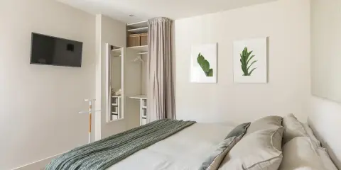 Ein offener Kleiderschrank, der wie ein Kleiderschrank selber bauen mit Vorhang wirkt, mit Regalen und Kleiderstange. Daneben ein Spiegel und ein Bett mit grüner Decke.