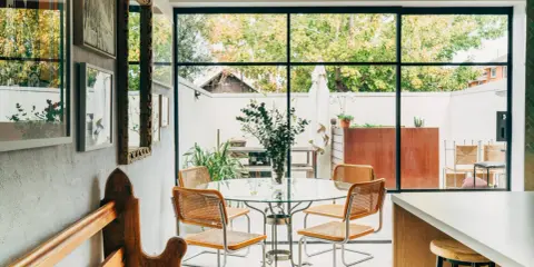 Sedie in paglia e metallo, ispirate a Marcel Breuer, attorno a un tavolo di vetro in una luminosa sala da pranzo con vista sul giardino.