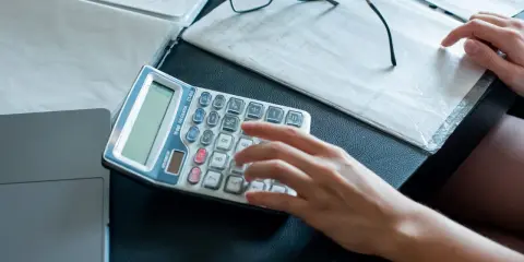 Ręce liczą na kalkulatorze dokumenty, analizując umowę zlecenie a alimenty. Obok leżą okulary i notatki.