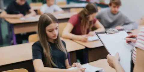 Leerlingen in een klaslokaal, sommigen schrijven in hun schrift. De leraar houdt een clipboard vast, misschien om te zien wat zijn leerlingen leren over wat zijn beta vakken.