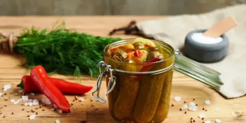Słoik pełen pikantnych ogórków po meksykańsku, obok świeży koperek i czerwone papryczki chili.