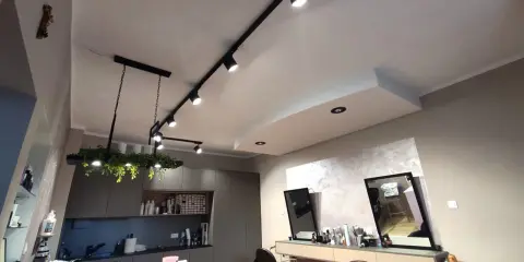 Nowoczesne oświetlenie w salonie fryzjerskim tworzy przytulną atmosferę. Reflektory na szynach i wisząca lampa z zielenią podkreślają styl wnętrza.