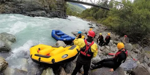 Grupa w kamizelkach ratunkowych i kaskach przygotowuje się do raftingu na rwących rzekach w Austrii. Dwie dmuchane łodzie czekają na brzegu.
