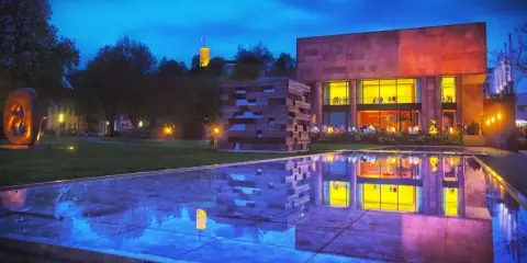 Abendstimmung am Museum mit reflektierendem Wasserbecken. Ein Turm leuchtet in der Ferne. Entdecke Bielefeld Geheimtipps!