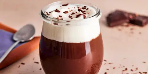 Ein Glas mit Omas Schokoladenpudding mit Sahne und Schokostreuseln. Daneben liegen ein Löffel und ein Stück Schokolade.