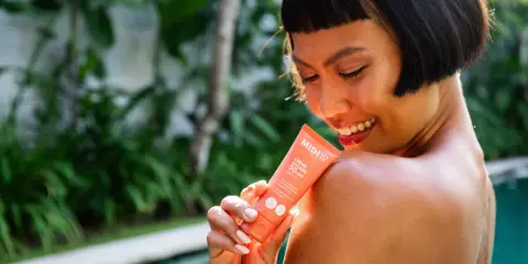 Une femme souriante applique une crème solaire SPF 50+ sur son épaule. Le tube indique "Très haute protection", le SPF 50 signification est une protection optimale.