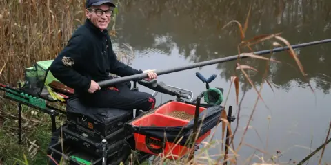 Wędkarz z uśmiechem na twarzy, przygotowany do łowienia metodą feeder. Widać jego sprzęt i zanętę w czerwonych pojemnikach.