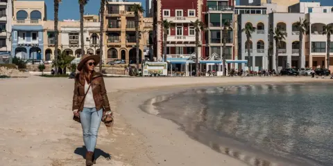 Kobieta spaceruje po piaszczystej plaży w pretty bay Malta, w tle kolorowe budynki i palmy.
