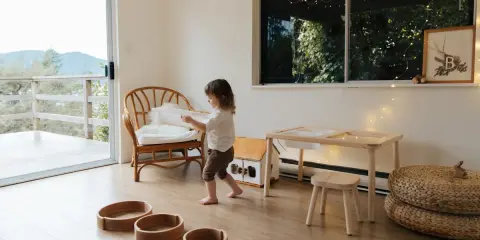 Bambina gioca in una stanza giochi in casa, tra sedie di vimini, tavolino e cesti.