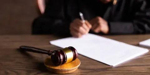 Młotek sędziowski na drewnianym stole, w tle osoba pisząca na dokumencie.