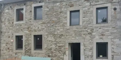 Maison ancienne en cours de rénovation, avec des murs en pierre brute et de nouvelles fenêtres. Le projet de construire en pierre continue.