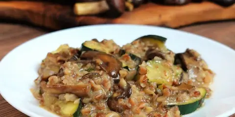 Pyszne kaszotto z grzybami i cukinią na białym talerzu, w tle drewniana deska z grzybami.