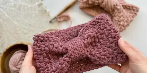 Manos sostienen una diadema tejida a crochet con un lazo. Detrás, otra diadema y ovillo de lana. ¡Descubre patrones gratis de diademas de ganchillo!