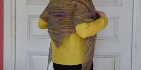 En person visar upp en vackert stickad halsduk i mosstickning med färgskiftningar i gult och lila.