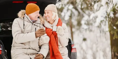 Ett äldre par sitter i bakluckan på en bil i snöig skog, njuter av livet med varma drycker.