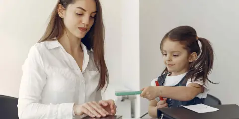 Pedagog specjalny wspiera dziecko w nauce. Kobieta i dziewczynka siedzą przy stole, rysują.