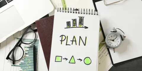Na biurku leżą dokumenty, laptop, okulary i budzik. Na notatniku widnieje napis "PLAN" i schemat, który symbolizuje indywidualny plan rozwoju.
