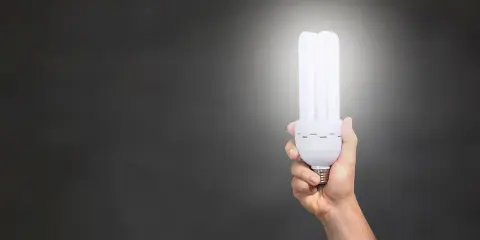 Dłoń trzymająca energooszczędną żarówkę o mocy 806 lumenów, rozświetlającą ciemne tło.