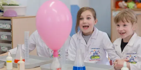 Dzieci w fartuchach laboratoryjnych z ekscytacją obserwują dmuchany balon podczas eksperymentu. To świetna chemia dla dzieci!